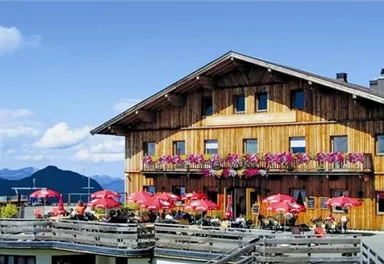 Ein rustikales Holzhaus mit Balkonen, die mit Blumen geschmückt sind. Im Vordergrund stehen Tische mit roten Sonnenschirmen, umgeben von einer malerischen Berglandschaft.