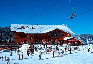 Ein Skigebiet mit vielen Menschen, die Ski fahren und spielen. Im Hintergrund ist ein rotes Gebäude und ein blauer Himmel sichtbar.