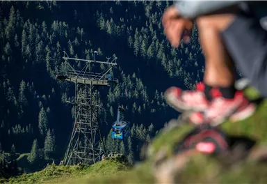 Eine Seilbahn fährt über eine grüne Berglandschaft. Im Vordergrund befindet sich eine Person, die auf dem Boden sitzt.