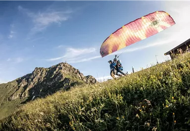 Ein Paraglider läuft auf einer Wiese mit Bergen im Hintergrund. Der Himmel ist blau und die Landschaft ist grün und sonnig.