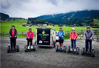 Eine Gruppe von fünf Personen steht auf Segways in einer malerischen Landschaft. Im Hintergrund sind grüne Wiesen und Berge unter einem bewölkten Himmel zu sehen.