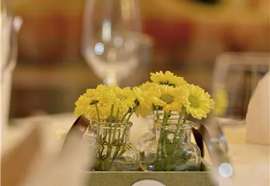 A table setting with a small flower vase and yellow flowers. In the background, glasses and a subtle atmosphere can be seen.