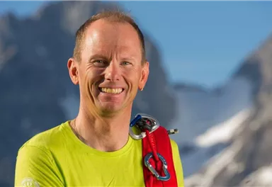 A smiling man in a bright yellow shirt stands in front of a mountainous landscape. He is wearing climbing gear and radiates a sense of adventure.