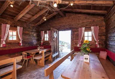 A cozy cabin with wooden walls and large windows. Inside are wooden tables and benches, as well as decorative curtains and a bouquet of flowers.