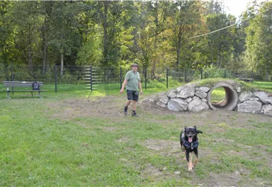 A man is walking in a dog park. A dog is happily running towards him.