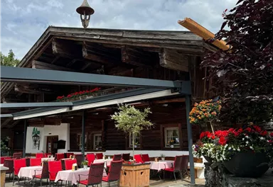 A cozy restaurant with a wooden facade and red tablecloths.  
Surrounded by flowers and outdoor seating under a cloudy sky.