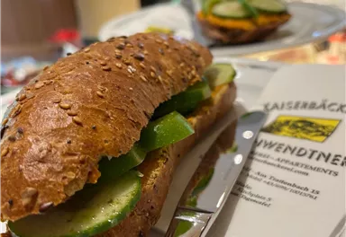 A delicious sandwich on a plate, filled with cucumbers and green peppers. In the background, more snacks and an open environment can be seen.
