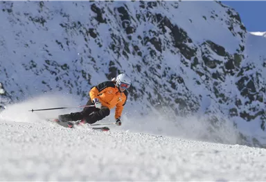Ein Skifahrer in oranger Jacke fährt schnell über einen schneebedeckten Hang. Im Hintergrund sind beeindruckende Berge zu sehen.