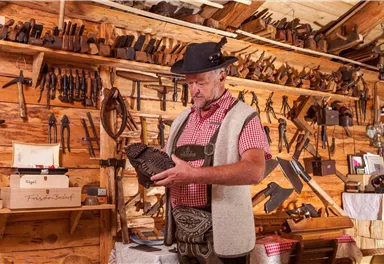 A craftsman in traditional clothing is examining an intricately carved wooden piece. In the background, many tools are visible on a wooden wall.