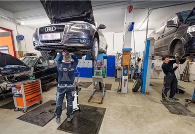 In a workshop, two cars are held by lifts. Mechanics are working on the vehicles and performing repairs.