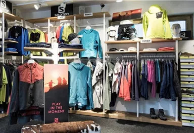 Ein Geschäft für Sportbekleidung mit einer Vielzahl von Jacken, T-Shirts und Schuhen. Im Vordergrund steht ein Werbeschild mit dem Slogan "PLAY IN THE MOMENT".