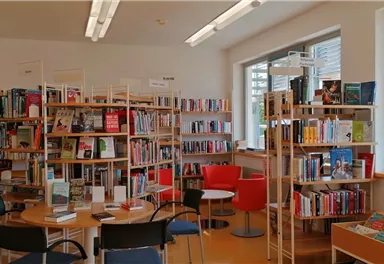 A bright library with shelves full of books. In the middle are tables and chairs for readers.