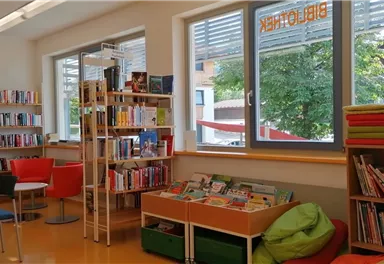 A bright room with bookshelves and cozy seating. A view of the trees outside is visible through the windows.
