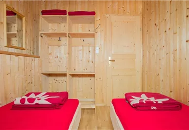 A cozy room with two single beds and red towels. The walls are made of light wood, and there is a mirror as well as a wardrobe.