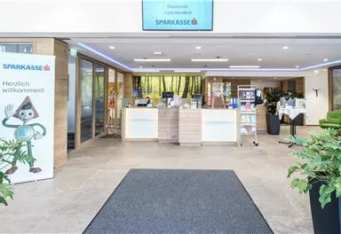 A bright and inviting bank branch with a reception area. In the foreground, there is a carpet, and there are plants as well as information stands.