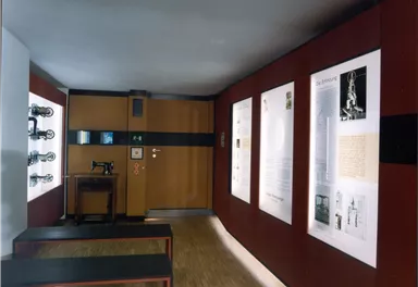 Ein Ausstellungsraum mit Wänden, die mit Informationsplakaten und Bildern dekoriert sind. In der Mitte stehen zwei lange Bänke.