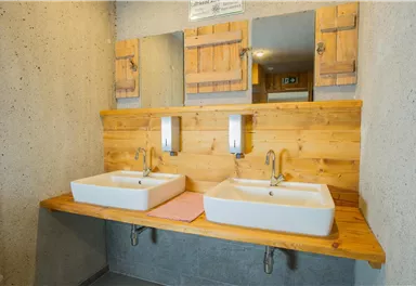 A modern bathroom with two sinks on a wooden shelf. In the background, mirrors and rustic wooden contours can be seen.