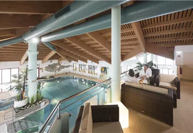 An elegant indoor pool with a wooden ceiling and plants. In the foreground, two people are sitting on a cozy seating group.
