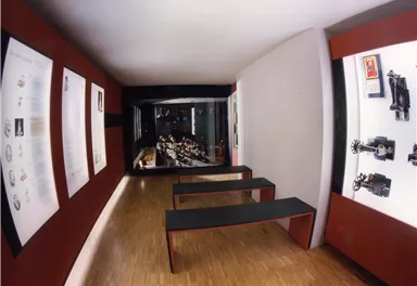 Ein moderner Ausstellungsraum mit Ausstellungsstücken an den Wänden und Sitzgelegenheiten in der Mitte. Die Beleuchtung ist gedämpft und schafft eine gemütliche Atmosphäre.