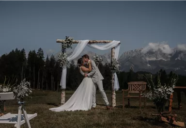 A bridal couple kisses in front of a festively decorated wedding arch in a picturesque mountain landscape. The scene is surrounded by lush greenery and mountains.