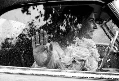 A bride is sitting in the car and has her hand against the window. It is raining outside as she gazes lovingly into the distance.