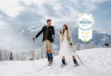 A bridal couple stands in a snowy mountain landscape, looking at each other. They are wearing winter clothing and ski equipment, surrounded by snow-covered trees and mountains.