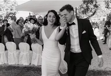A happy bride and groom leave the ceremony while the guests applaud. The bride wears an elegant white dress and holds a bouquet in her hand.