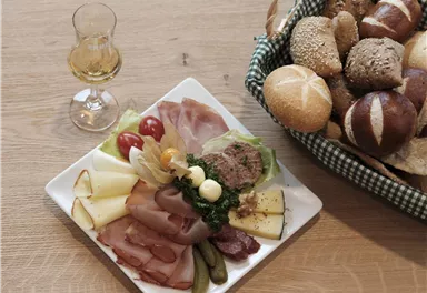 Ein Teller mit verschiedenen Wurst- und Käsesorten, eingelegten Gurken und Kirschtomaten. Daneben steht ein Glas und ein Korb mit Brötchen.