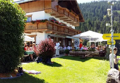 Ein gemütliches Restaurant mit Sonnenschirmen und Gästen im Freien, umgeben von grünen Wiesen und Bergen. Die Fassade des Hauses ist im traditionellen Stil mit Blumenkästen dekoriert.