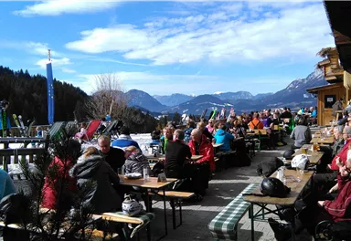 Eine belebte Terrasse mit vielen Gästen, die die Sonne genießen. Im Hintergrund sind Berge und ein klarer Himmel zu sehen.