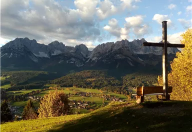 Eine malerische Berglandschaft mit hohen Gipfeln und buntem Herbstwald. Im Vordergrund steht ein Holzbank mit einem Kreuz, das zur Aussicht einlädt.