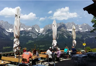 Eine schöne Berglandschaft mit schneebedeckten Gipfeln und einem klaren Himmel. Auf einer Terrasse sitzen Gäste und genießen die Aussicht.