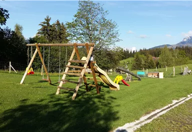 Ein schöner Spielplatz mit einer Schaukel und einer Rutsche. Im Hintergrund sind grüne Wiesen und Berge zu sehen.