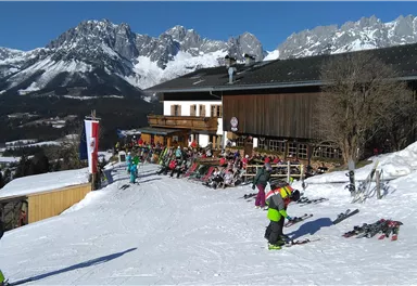 An alpine ski landscape with snow-covered mountains and a wooden house in the background. Several skiers and snowboarders are enjoying the slope.