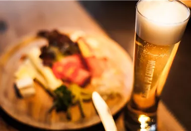 Ein Glas Bier steht neben einem Teller mit verschiedenen Wurst- und Käsesorten. Die Szene ist gemütlich und einladend.