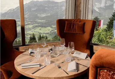 A stylishly furnished dining area with a round wooden table and cozy, high chairs. In the background, an impressive mountain landscape can be seen.
