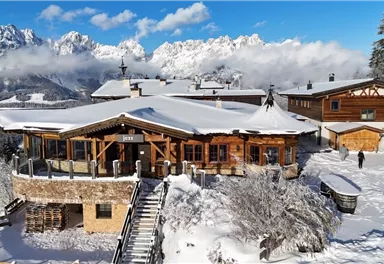 A rustic mountain cabin in the snow with a stunning view of the surrounding mountains. The sky is clear and the landscape is characterized by a beautiful winter atmosphere.