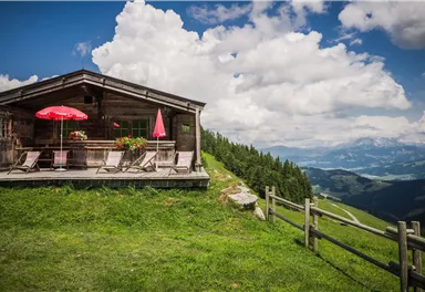 A cozy alpine cabin on a green meadow. In the background, clouds and mountains can be seen.