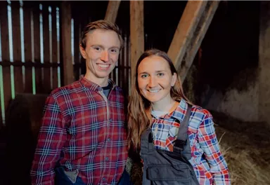 Ein junges Paar steht in einem Stall. Beide tragen karierte Hemden und lächeln freundlich.