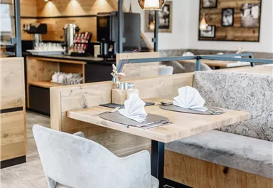 Ein modernes Restaurantinterieur mit Holz- und Grau-Tönen. Ein Tisch mit dekorierten Servietten und hellen Sitzplätzen lädt zum Essen ein.