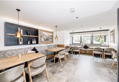 Ein modernes Restaurant mit hellen Wänden und Holzdeko. Es gibt gemütliche Sitzgelegenheiten und große Fenster mit Blick nach außen.