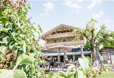 Ein malerisches Gebäude mit Balkon, umgeben von grünen Pflanzen und einem schönen Himmel. Im Vordergrund sind Tische und Stühle im Freien sichtbar.