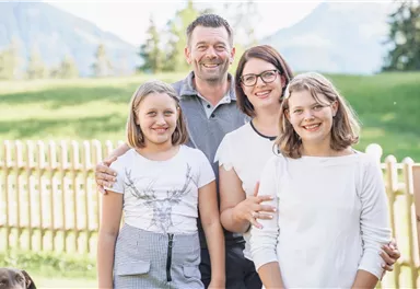 Eine fröhliche Familie posiert im Freien vor einer grünen Wiese und einem Zaun. Im Hintergrund sind Bäume und Berge sichtbar.