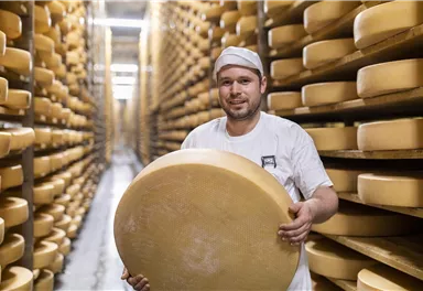 Ein Käsehersteller steht in einem Raum voller Regale mit Käselaiben. Er hält einen großen Käse in der Hand und lächelt.