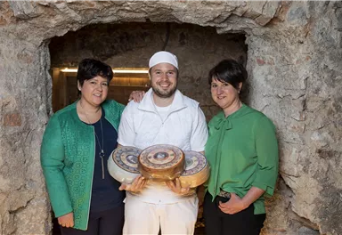 Drei Personen stehen vor einer Steinmauer. Der Mann in der Mitte hält Käseplatten, während die beiden Frauen an seiner Seite lächeln.