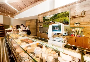 Ein einladendes Käsegeschäft mit Holzregalen und einer gläsernen Vitrine. Zwei Mitarbeiterinnen stehen hinter der Theke und bedienen Kunden.