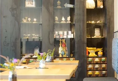 A modern café with bright wooden tables and cozy seating options. In the background, there are display cabinets with various decorative items.