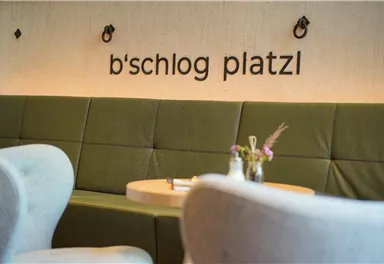 A cozy seating area with a green padded bench and modern chairs. The wall is decorated with the inscription "b'schlog platzl".