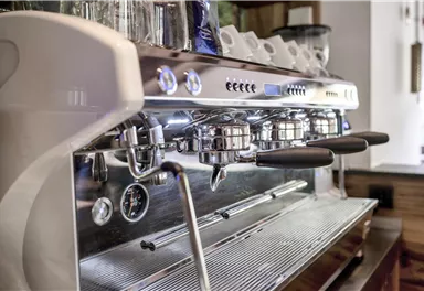 A modern espresso machine with shiny stainless steel and multiple brewing groups. The machine is positioned on a wooden countertop in a cozy café.
