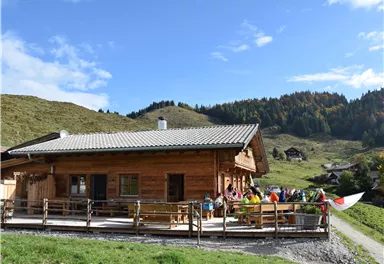 Eine gemütliche Berghütte umgeben von grünen Wiesen und Wald. Menschen genießen die sonnige Atmosphäre draußen auf der Terrasse.